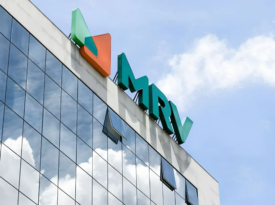 Logotipo da MRV no alto de um edifício com fachada de vidro que reflete o céu e as nuvens.