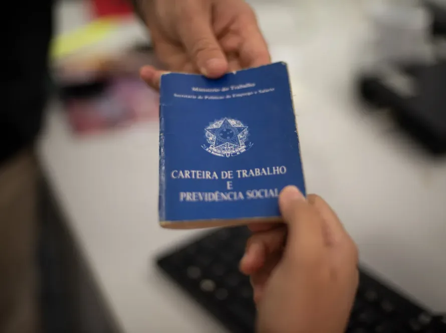 Carteira de Trabalho e Previdência Social sendo entregue de uma mão para outra sobre uma mesa de escritório