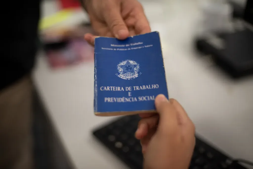 Carteira de Trabalho e Previdência Social sendo entregue de uma mão para outra sobre uma mesa de escritório